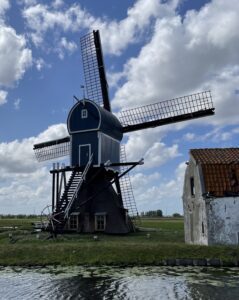 Een blauwe windmolen naast een oud gebouw in een groen landschap onder een bewolkte hemel.
