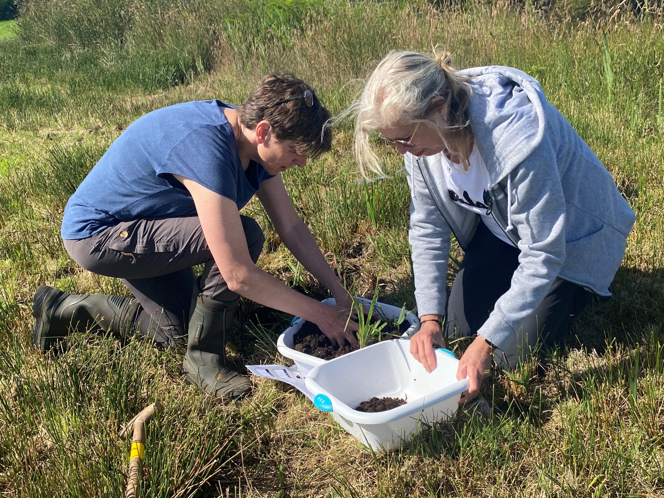 Twee mensen werken samen buiten, bezig met planten in bakken op grasland.