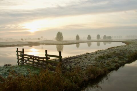 Mistige zonsopgang boven een rustig landschap met water, grasland en een houten hek op de voorgrond.