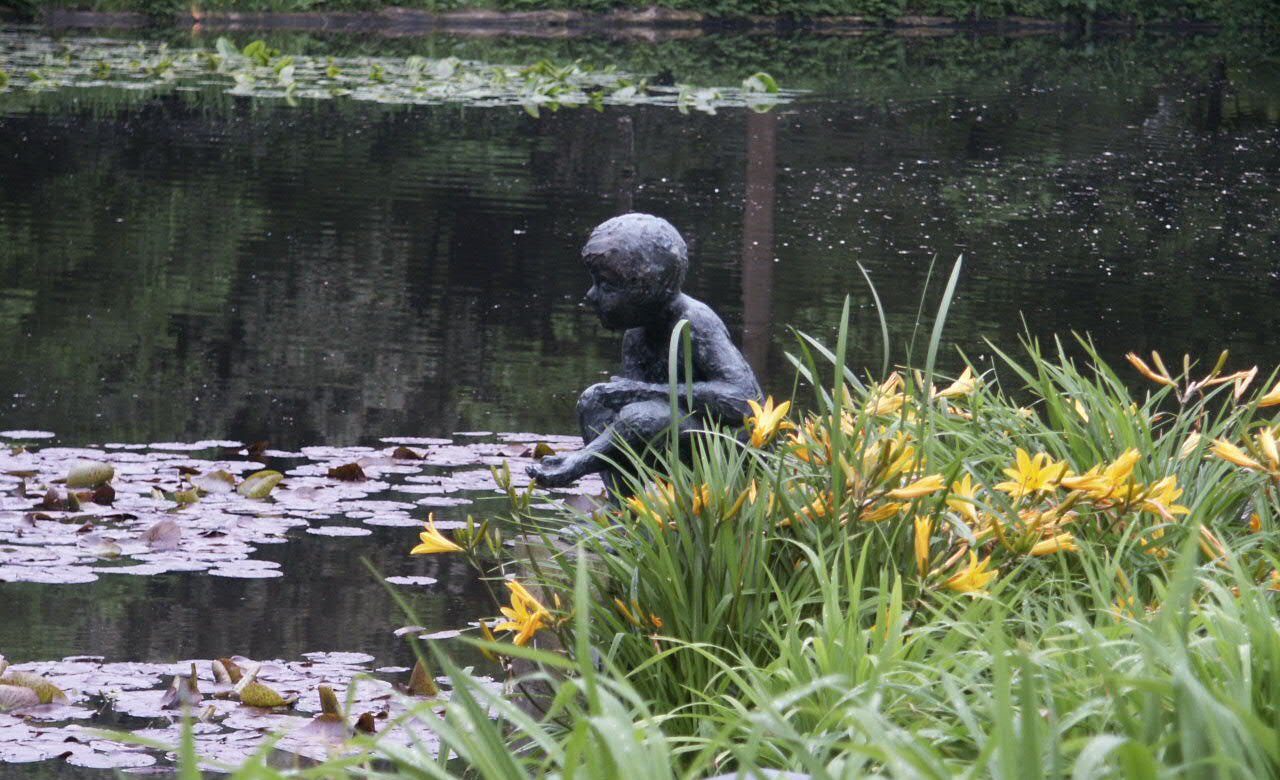 Standbeeld van een kind naast een vijver met waterlelies en omliggende gele bloemen.