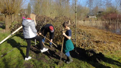 Drie mensen graven in een tuin naast een stilstaand water met riet, op een zonnige dag.