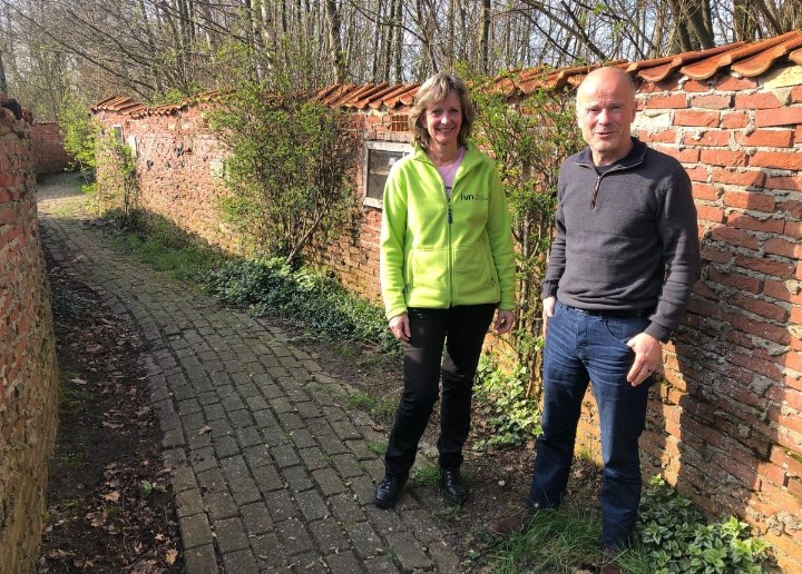 Twee mensen poseren op een stenen pad naast een bakstenen muur met planten.