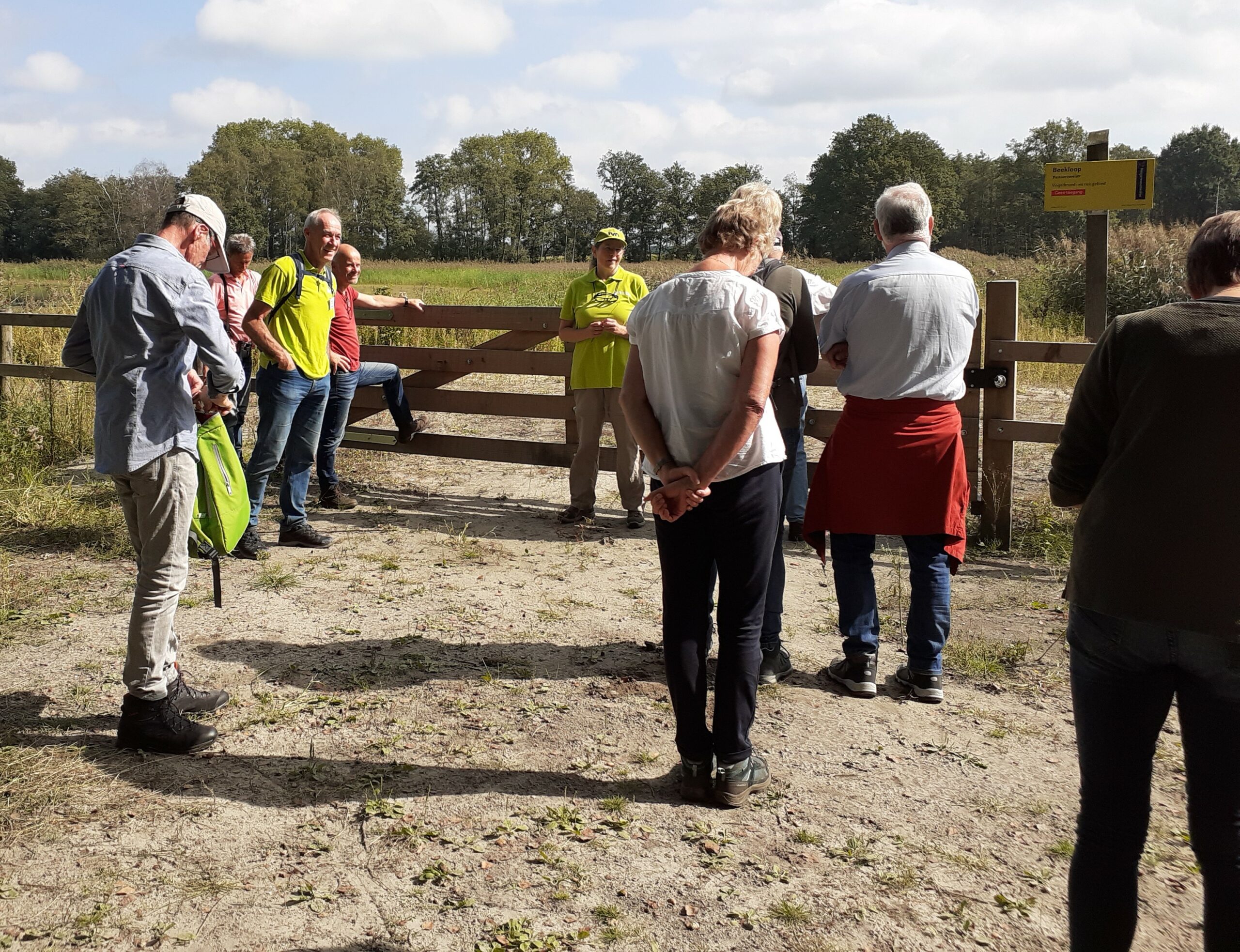 Groep mensen bij een hek tijdens een natuurwandeling, met bomen en een informatiebord op de achtergrond.