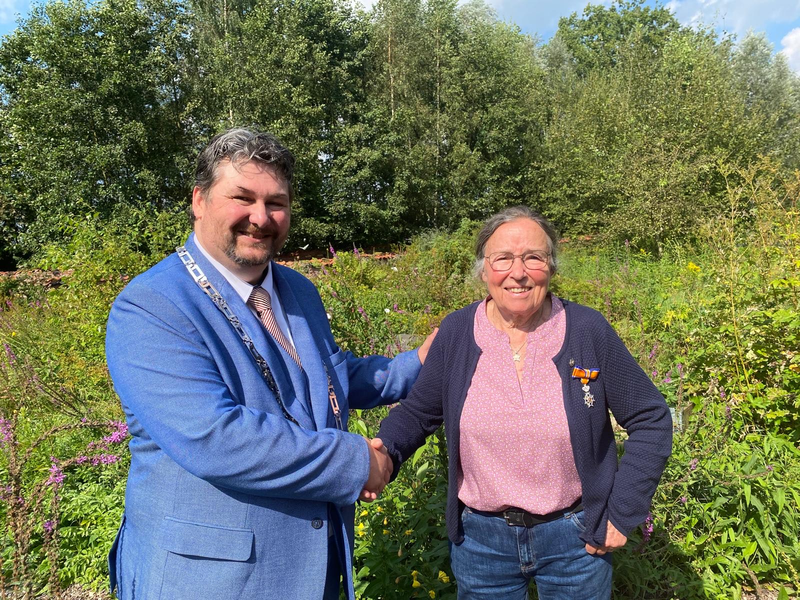 Twee mensen schudden handen in een groene tuin. Een persoon draagt een oranje lintje.