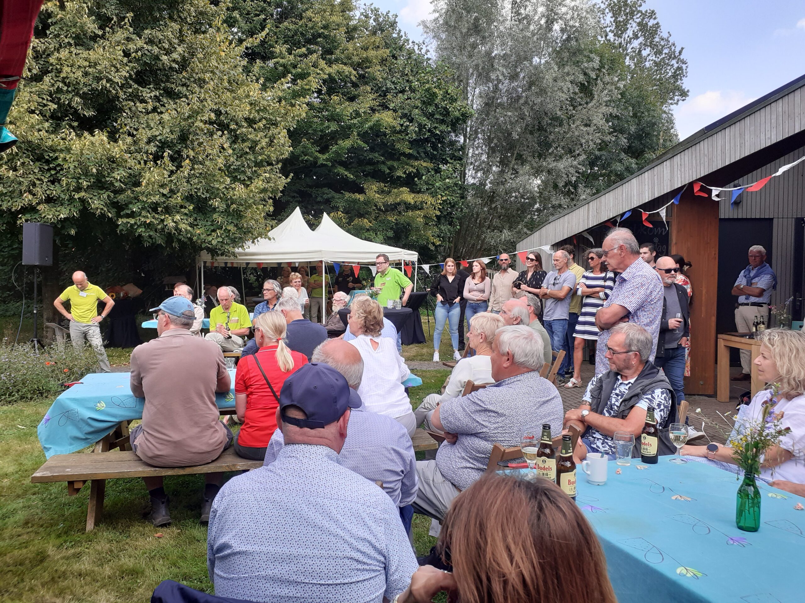 Groep mensen bij een buitenbijeenkomst met tafels, stoelen en een tent op een zonnige dag.
