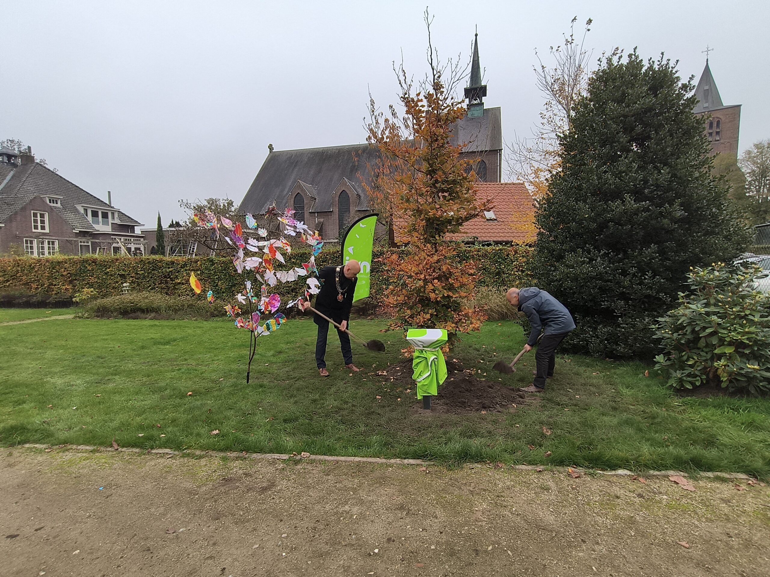 Twee personen planten een boom op een grasveld bij een kerk en een huis.