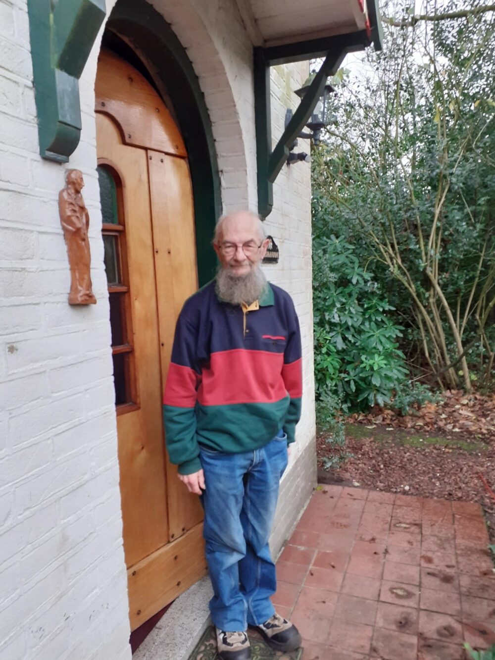 Een oudere man staat voor een houten deur, omringd door planten en een bakstenen muur.