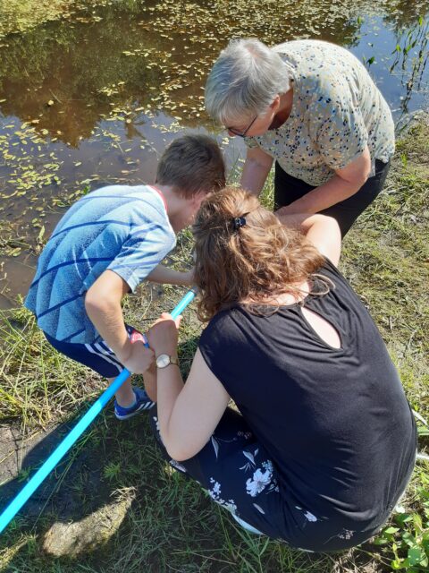 Drie mensen onderzoeken water bij een vijver met een blauwe stok op een zonnige dag.