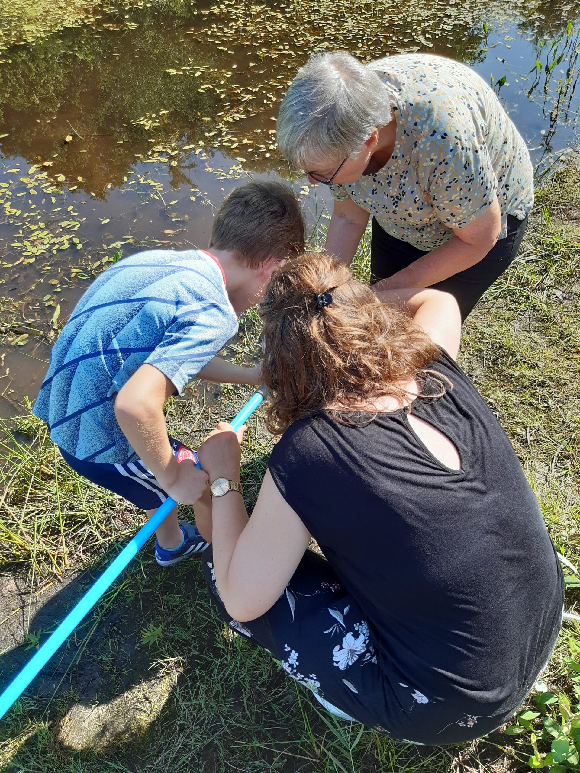 Drie mensen onderzoeken water bij een vijver met een blauwe stok op een zonnige dag.