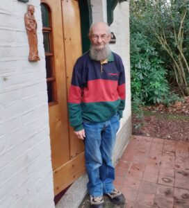 Een oudere man met een baard staat voor een houten deur, omgeven door groene struiken.