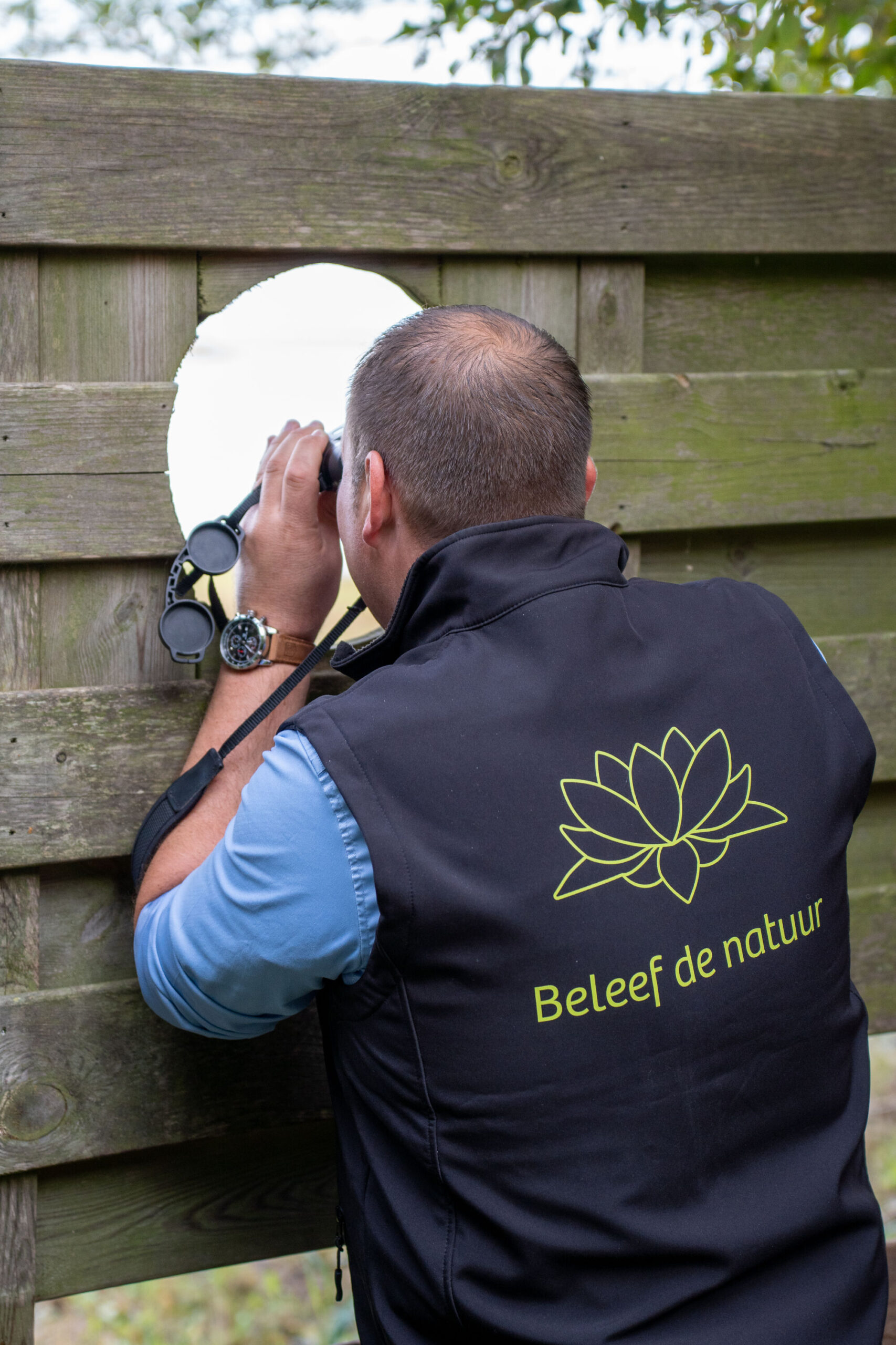 Man kijkt door een gat in een houten schutting met een verrekijker, vest met "Beleef de natuur".
