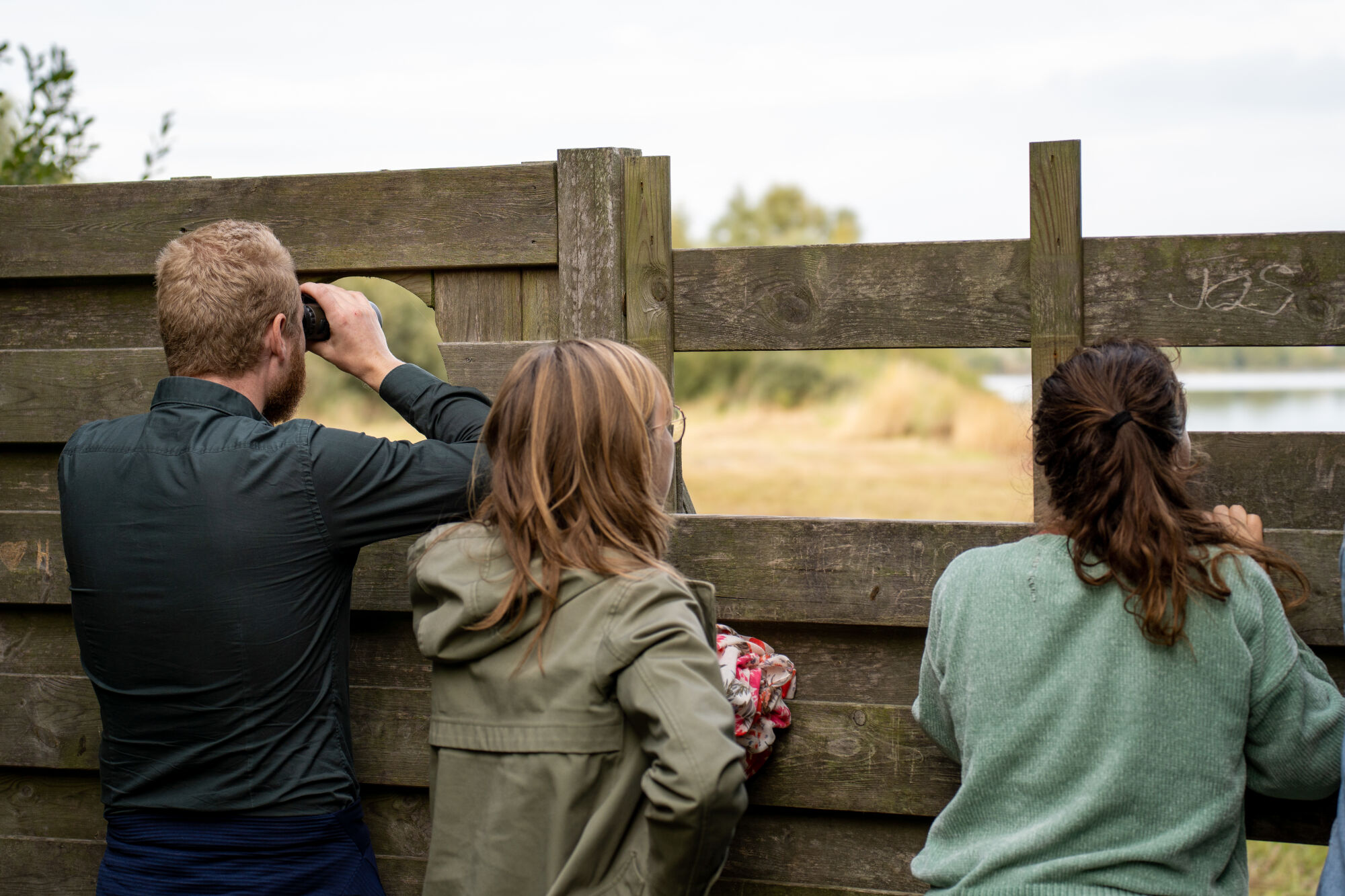 Drie mensen kijken door een houten kijkwand naar de natuur. Eén persoon gebruikt een verrekijker.