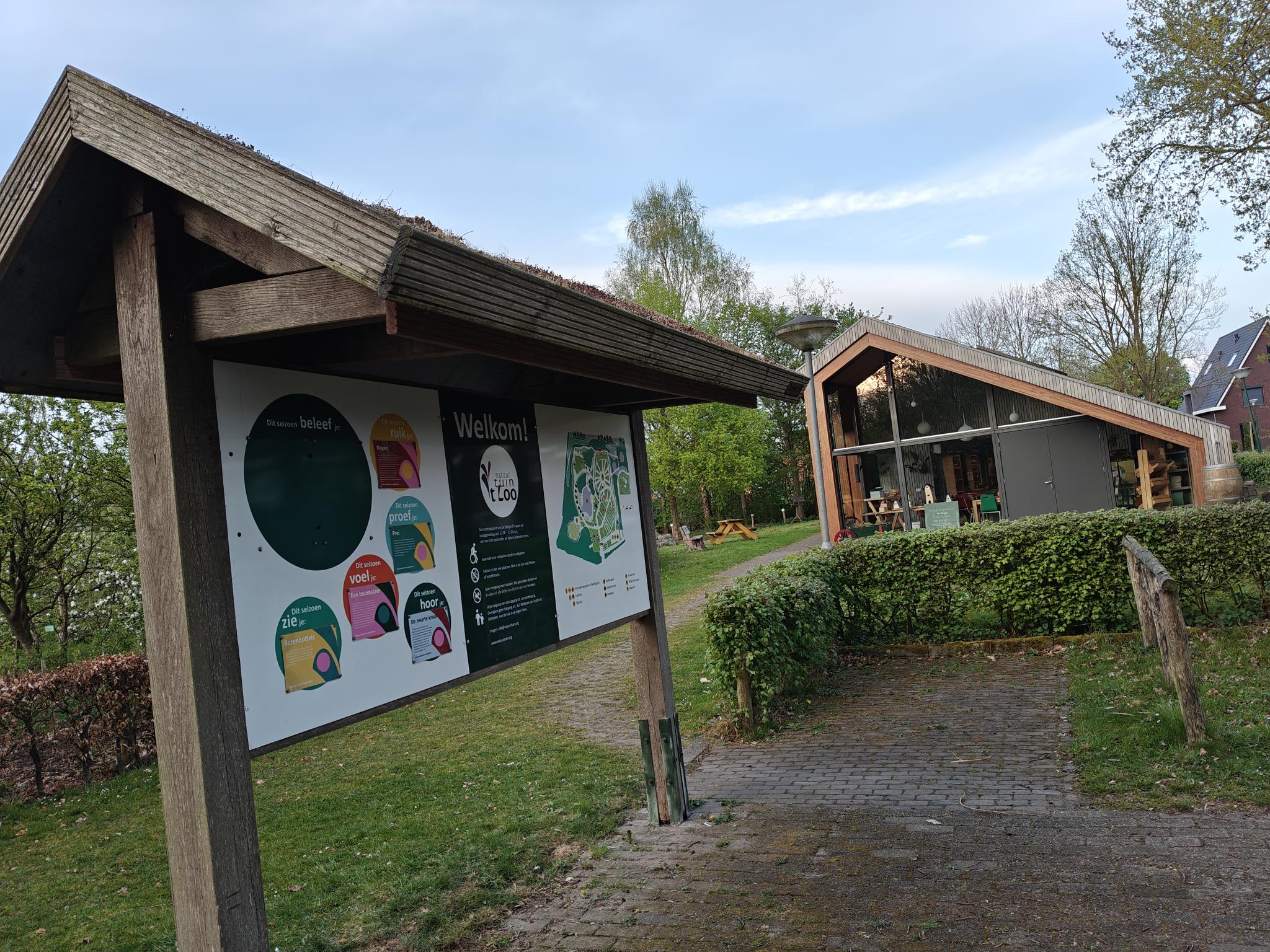 Infobord en modern gebouw in groene omgeving; uitnodigend welkom, met wandelpaden en bomen.