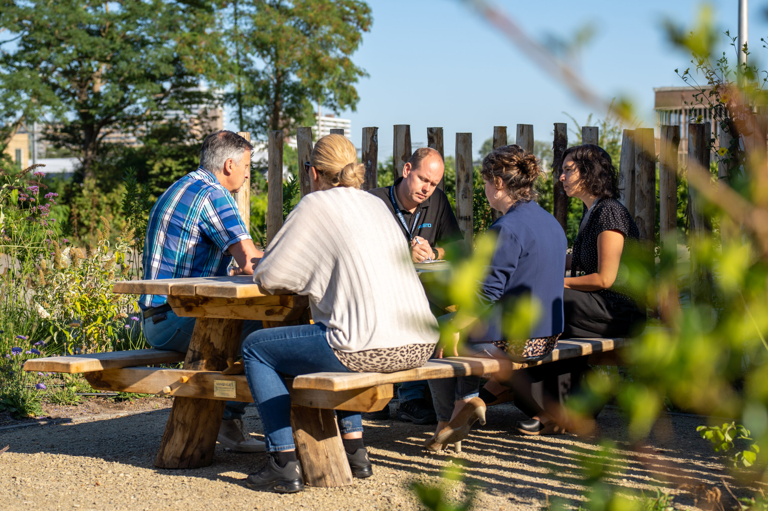 Vijf mensen vergaderen buiten aan een houten picknicktafel, omringd door groen.