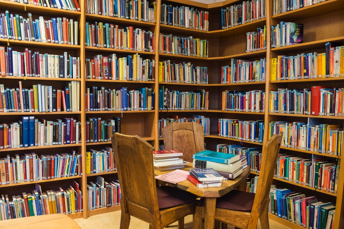 Bibliotheekhoek met gevulde boekenplanken, houten tafel en stoelen met stapels boeken.