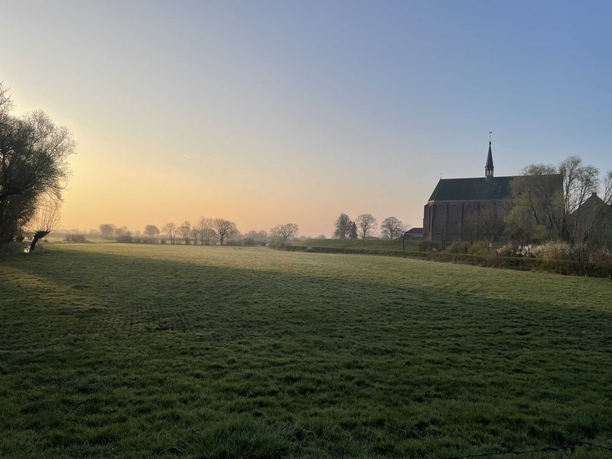 Landelijk landschap bij zonsopgang met een kerk aan de rechterkant en uitgestrekte grasvelden.