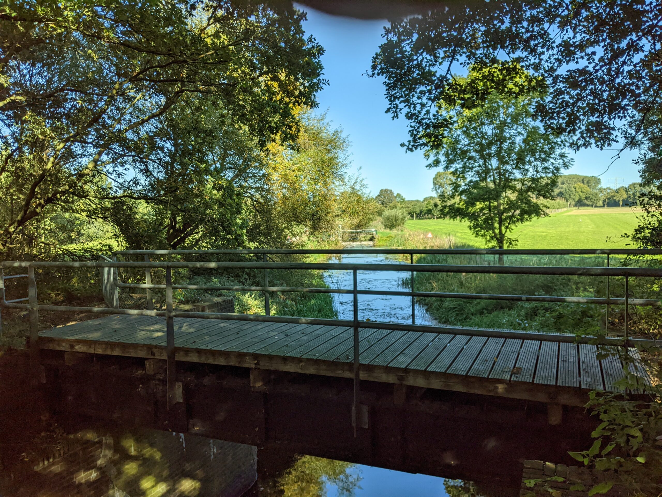 Houten brug over een smalle riviertje, omgeven door bomen en groene velden onder een blauwe hemel.