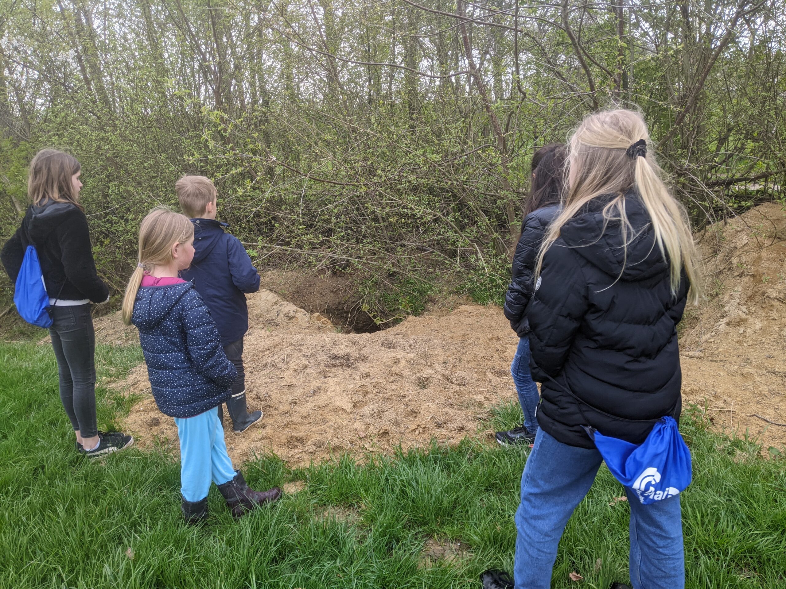 Groep kinderen kijkt naar een hol in de grond bij een bosrand.