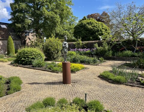 Tuin met grindpaden, groene struiken, paarse bloemen en een beeld op een sokkel.