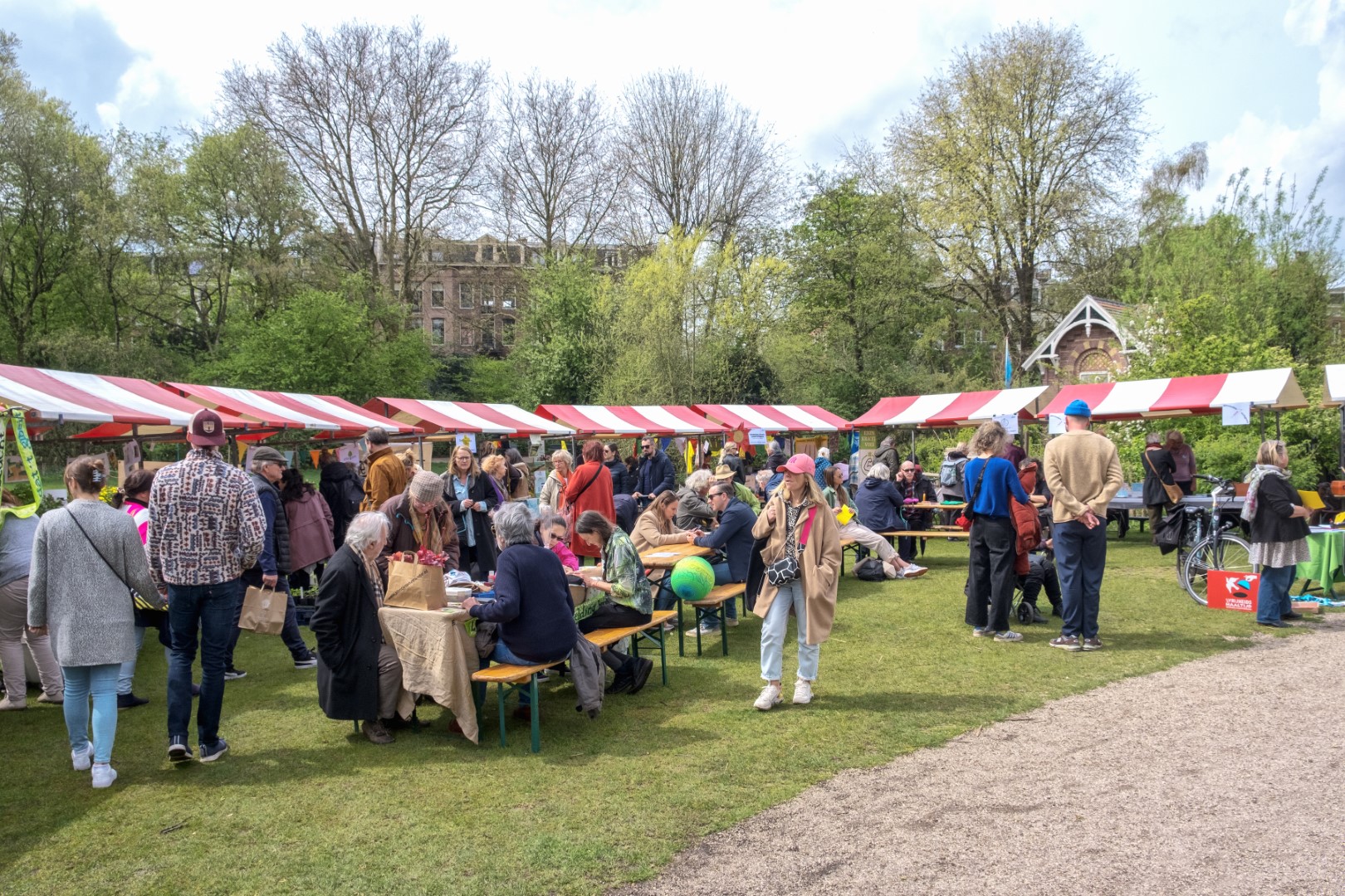 Buitenmarkt met mensen bij rood-witte kraampjes in een park met bomen.