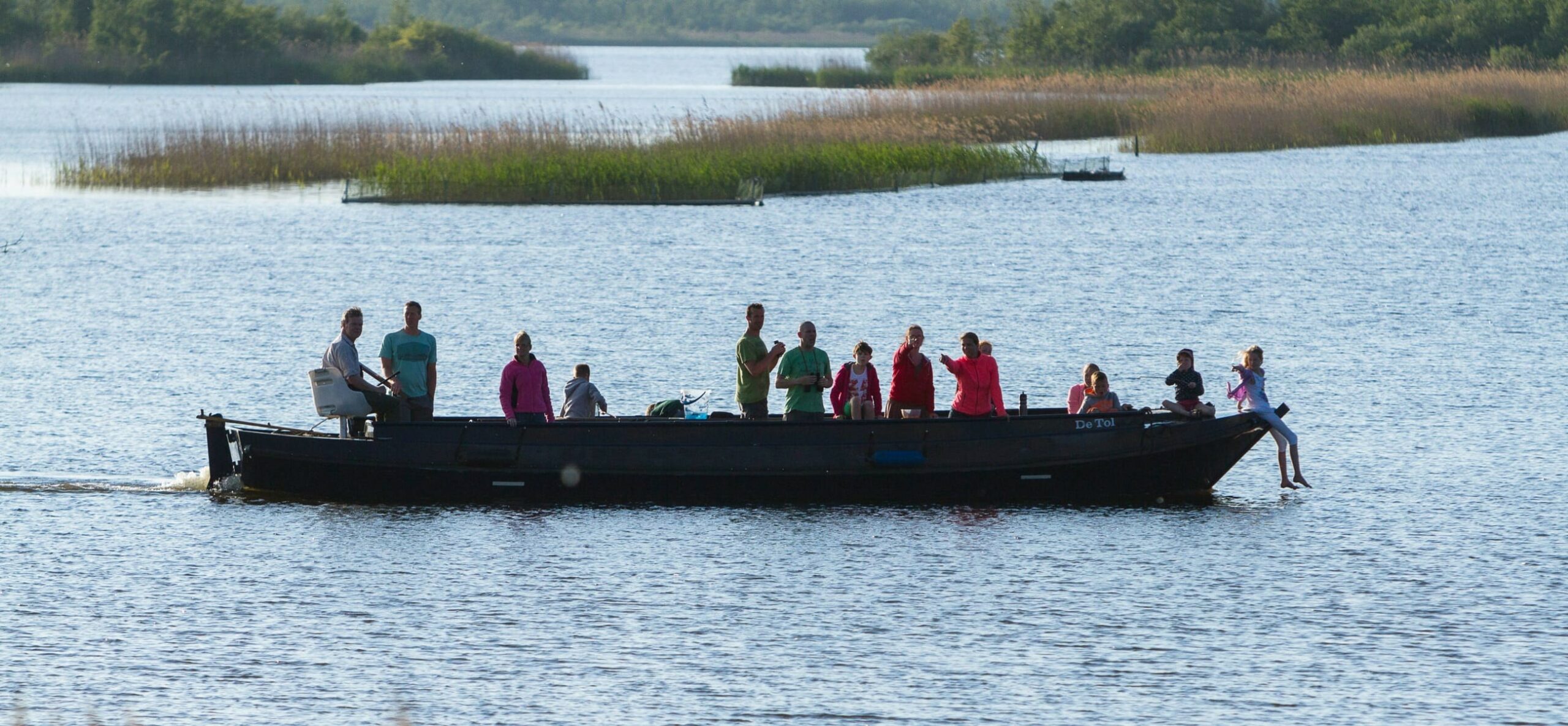 Boot met mensen op een meer, omringd door riet en bosrijk landschap op de achtergrond.