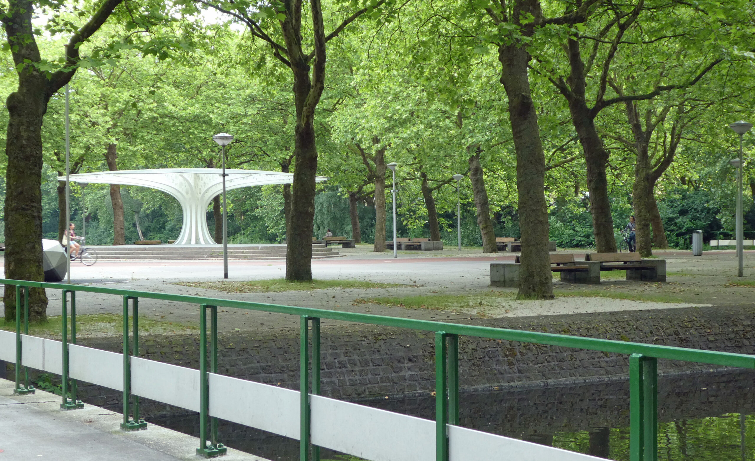 Park met bomen, banken en een witte, moderne overkapping. Vrouw fietst langs op een rustige dag.
