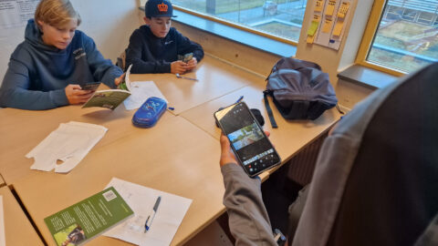 Drie kinderen werken aan een tafel; twee lezen, één kijkt naar foto's op een smartphone.