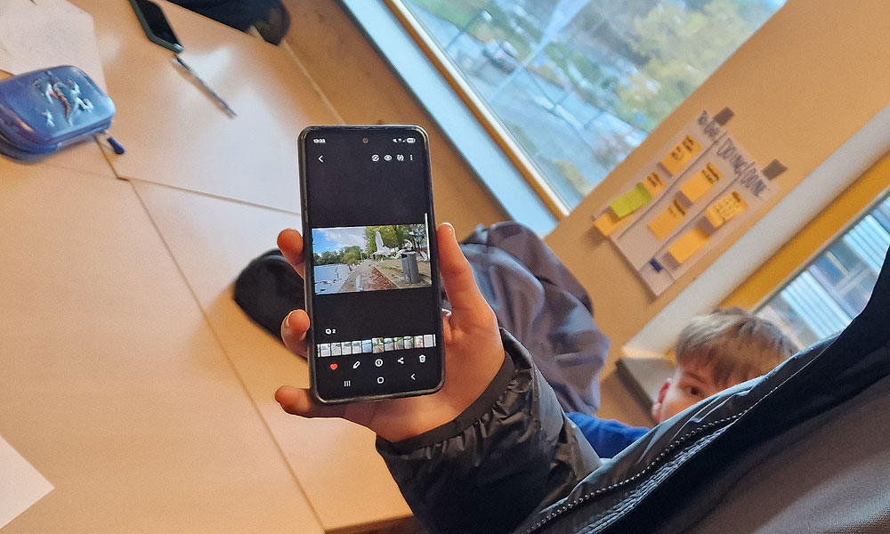 Persoon toont een foto op hun smartphone in een klaslokaal met raam op de achtergrond.