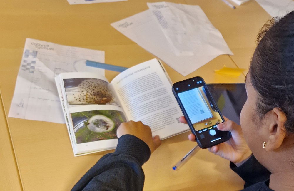 Persoon fotografeert een opengeslagen boek op tafel met een smartphone.