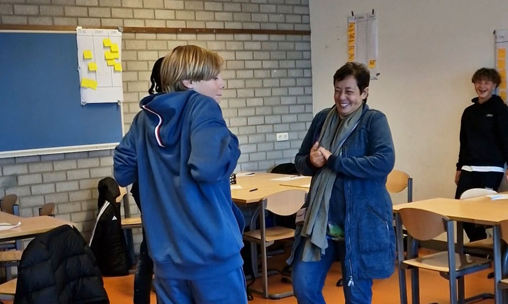 Groep mensen lachend in een klaslokaal met tafels en stoelen. Posters hangen aan de muur.