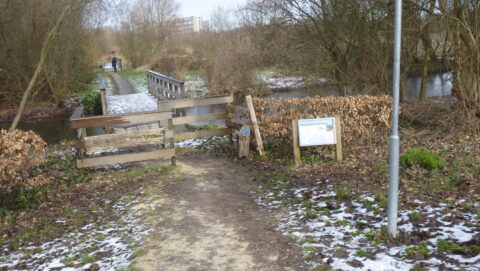 Ingang van een natuurpark met een houten poort, een pad en een besneeuwde brug over een beekje.