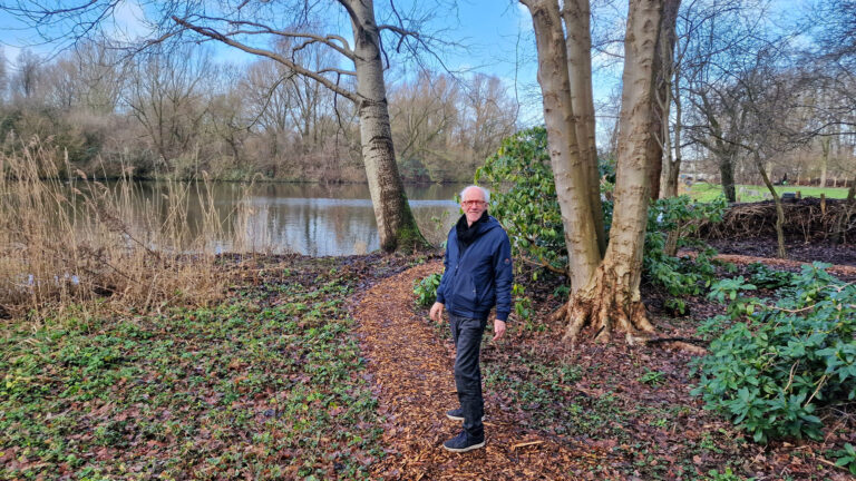 Persoon wandelt op bospaadje langs vijver met kale bomen en groene struiken.