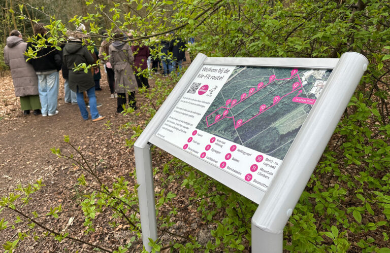 Groep mensen in een bos bij een informatiebord voor een fitnessroute, met bladeren op de voorgrond.