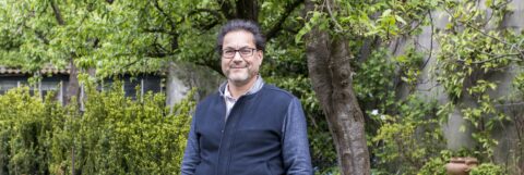 Man in donkere trui staat lachend in een groene tuin met bomen en struiken op de achtergrond.
