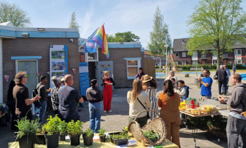 Mensen verzamelen zich buiten een gebouw met een regenboogvlag, etenstafel en planten.