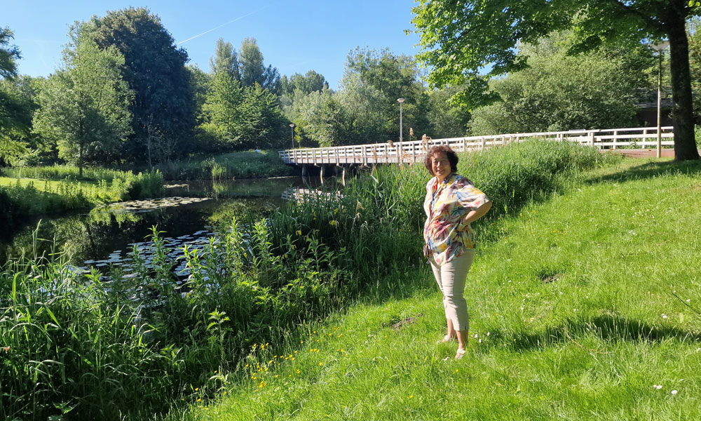 Vrouw poseert op grasveld bij rivier met lelies en houten brug onder blauwe lucht.