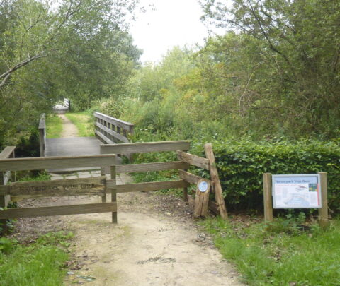 Pad en houten brug omgeven door groen met bord "Natuurpark Vrije Geer" aan de rechterkant.