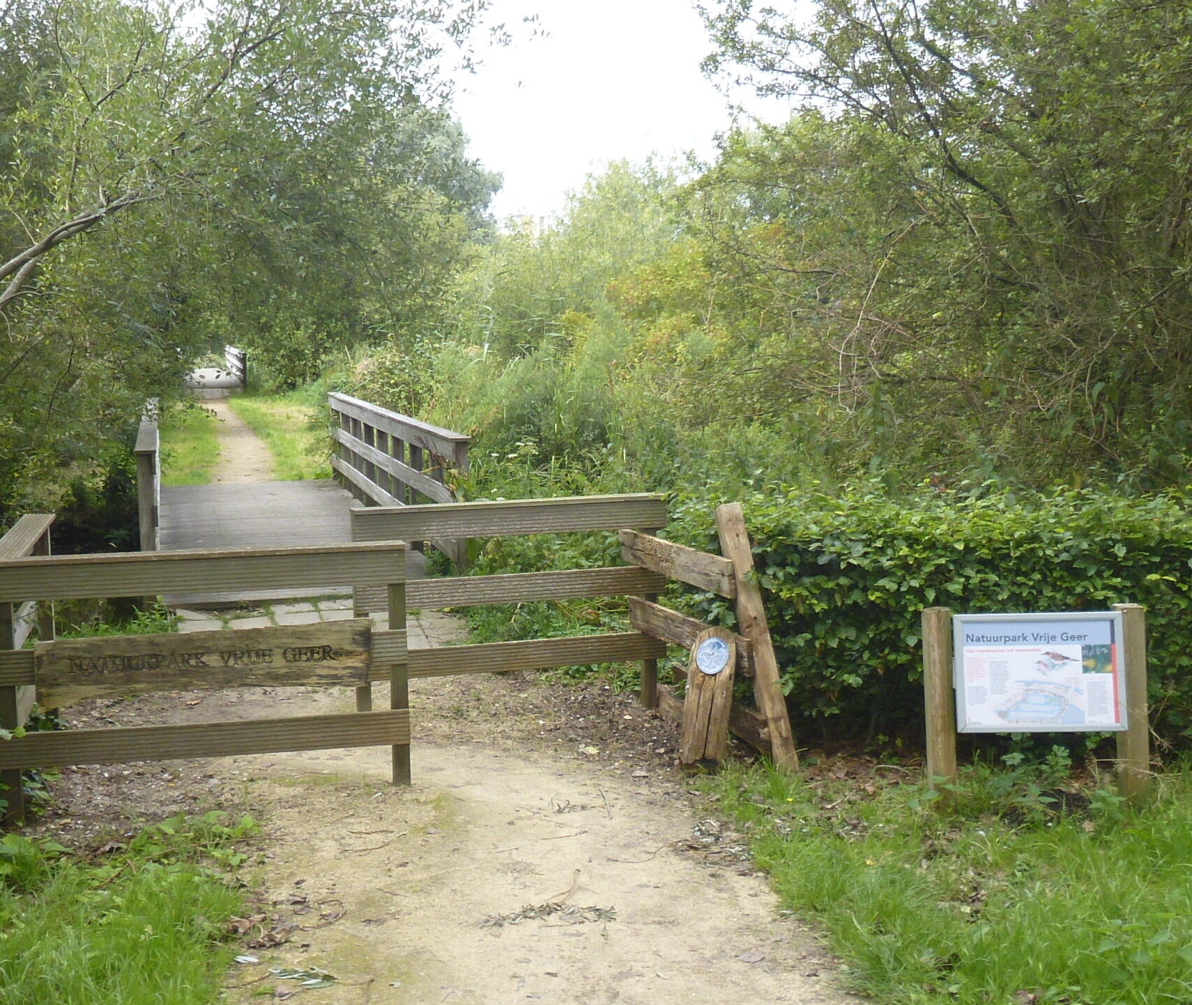 Pad en houten brug omgeven door groen met bord "Natuurpark Vrije Geer" aan de rechterkant.