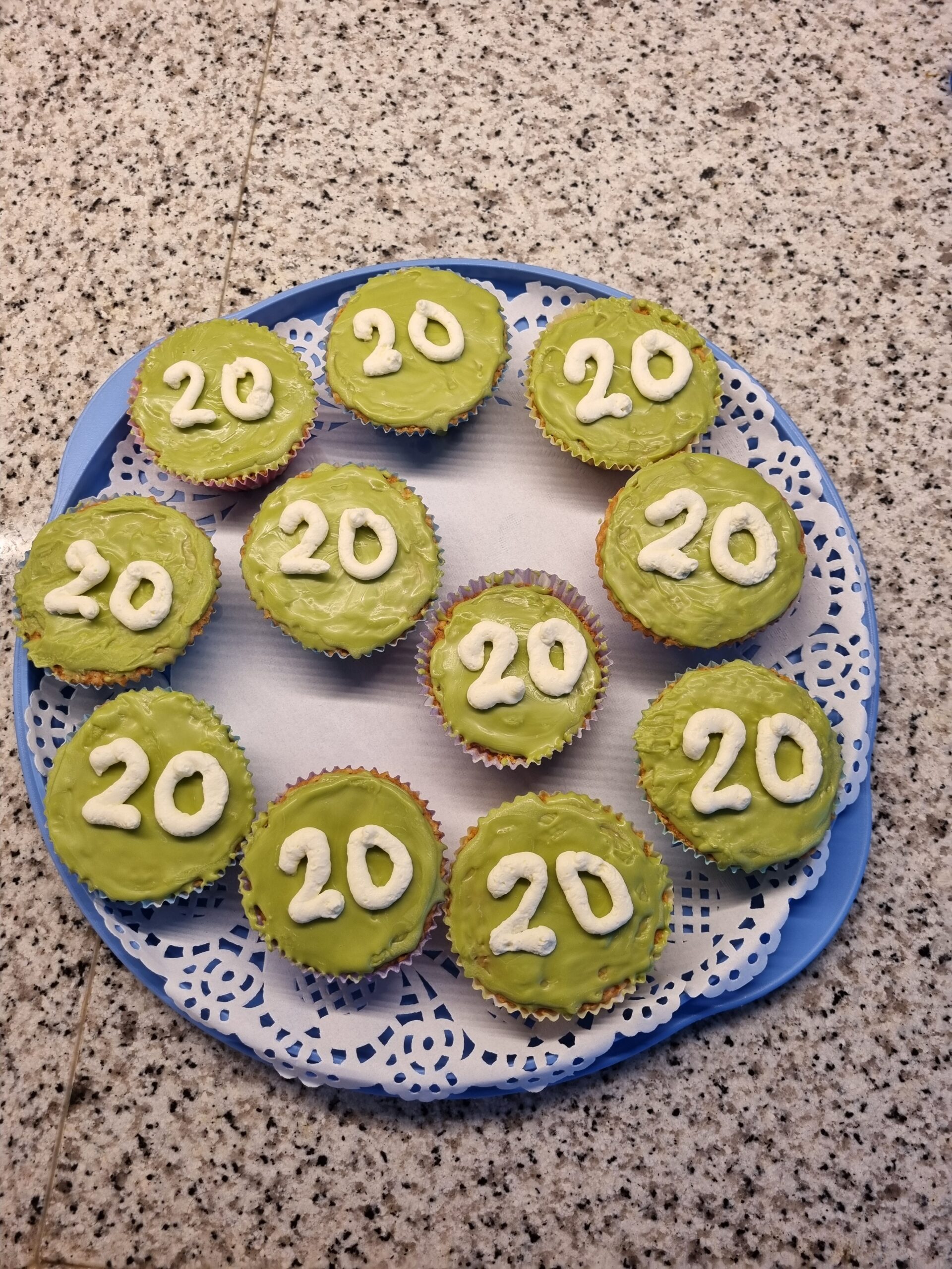 Elf cupcakes met groene icing en het cijfer "20" in wit, op een blauwe schaal met kanten onderlegger.