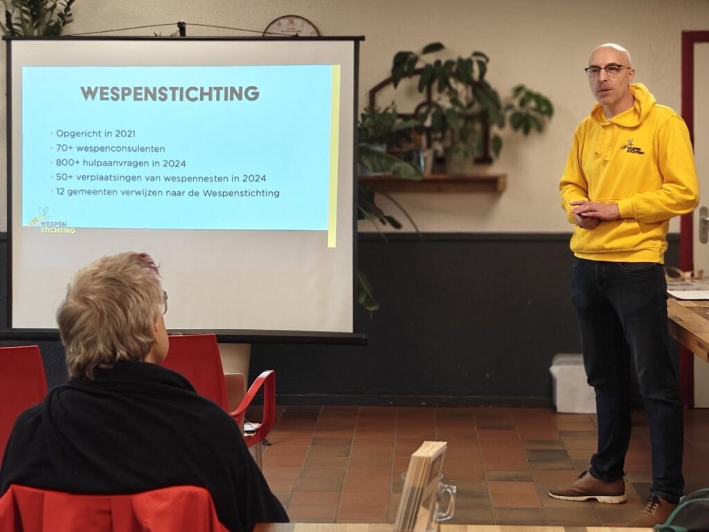 Man geeft presentatie over Wepenstichting voor publiek met dia op achtergrond.