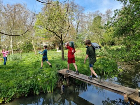 Kinderen dragen spullen over een houten brug in een groene, bosrijke omgeving.