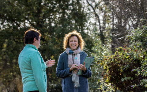 Twee vrouwen praten in een zonnige tuin, omgeven door groen en bloemen.