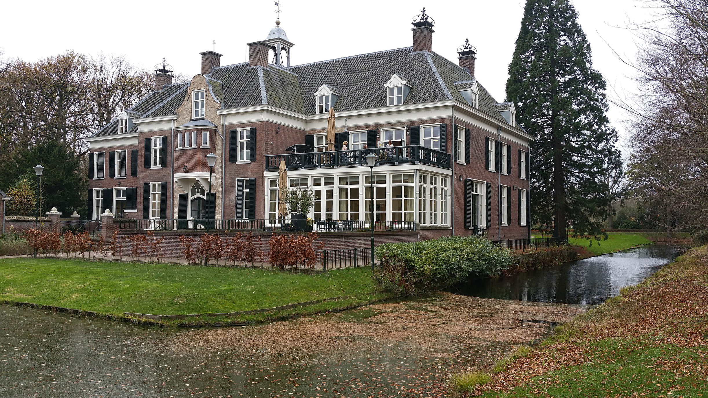 Een groot historisch herenhuis met een vijver en bomen in een herfstlandschap.