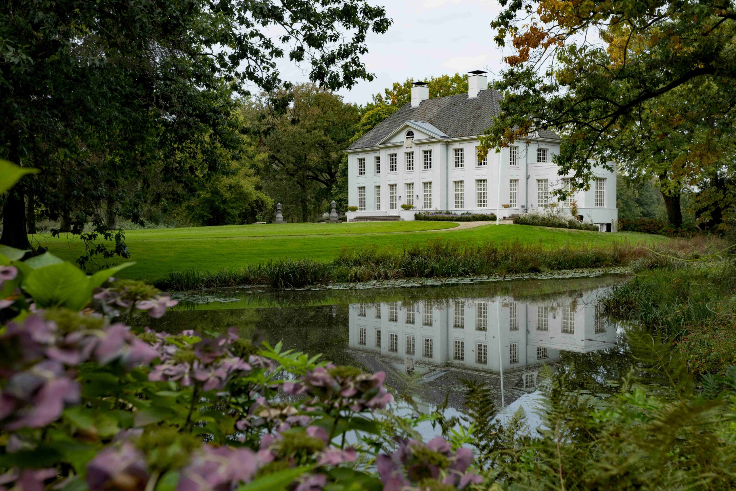 Wit landhuis weerspiegeld in een vijver, omgeven door groen en paarse bloemen.
