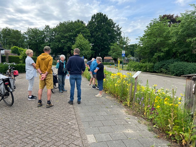 Groep mensen staan pratend op een stoep naast bloeiende planten en fietsen onder een bewolkte hemel.