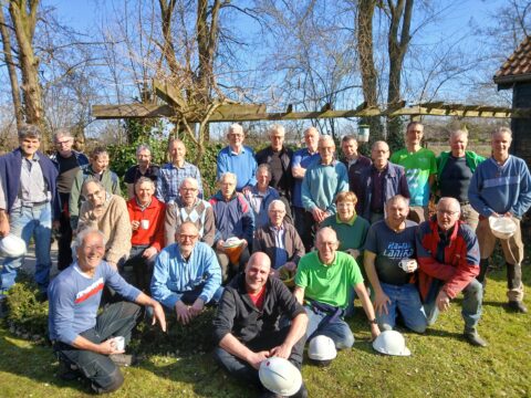 Groep mannen poseert buiten, sommigen met bouwhelmen, omgeven door bomen onder een heldere lucht.