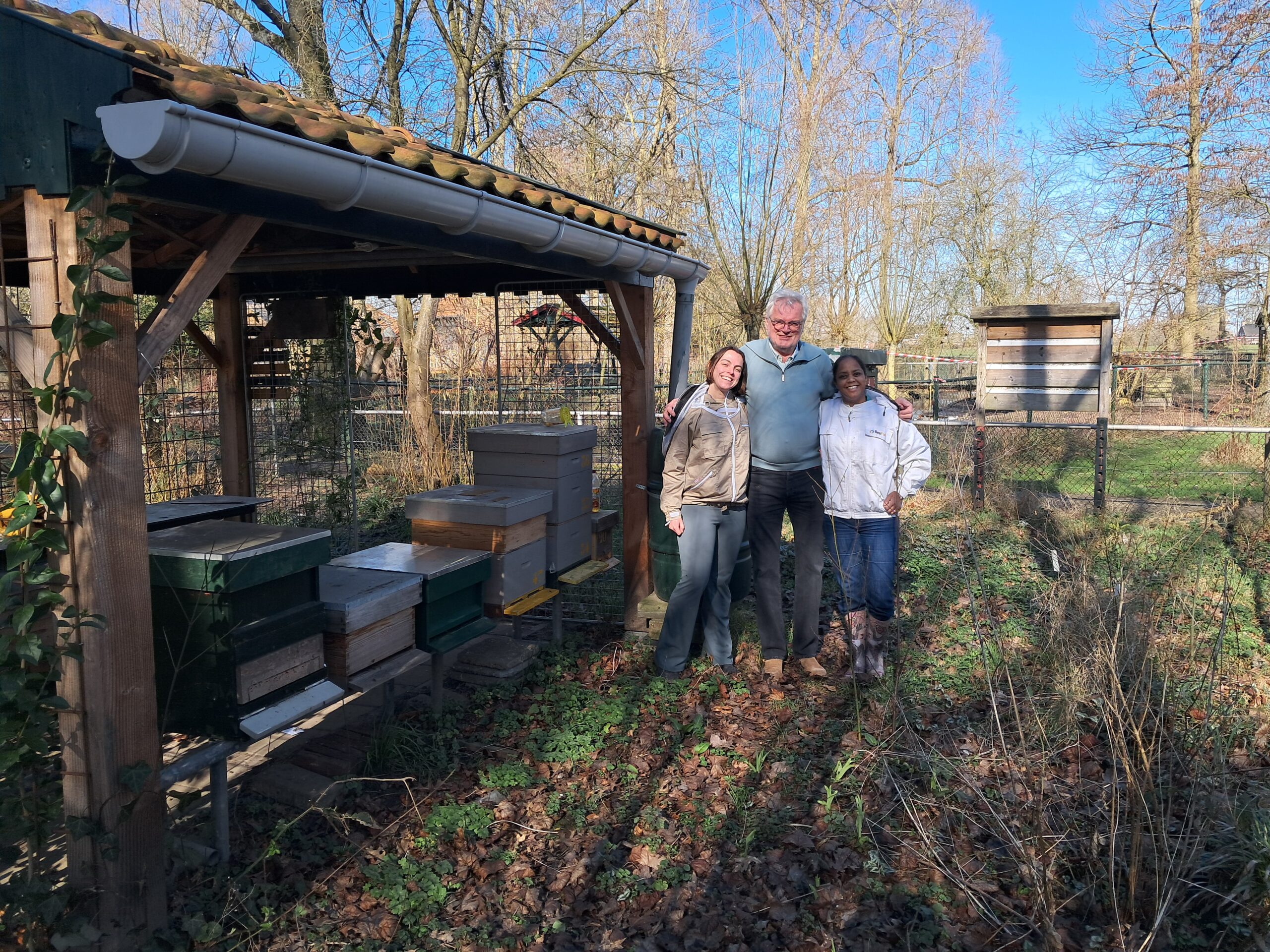 Drie mensen staan bij houten bijenkasten in een tuin, omringd door kale bomen.