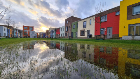 Rij kleurrijke huizen weerspiegeld in een grote plas, met bewolkte lucht en ondergaande zon.