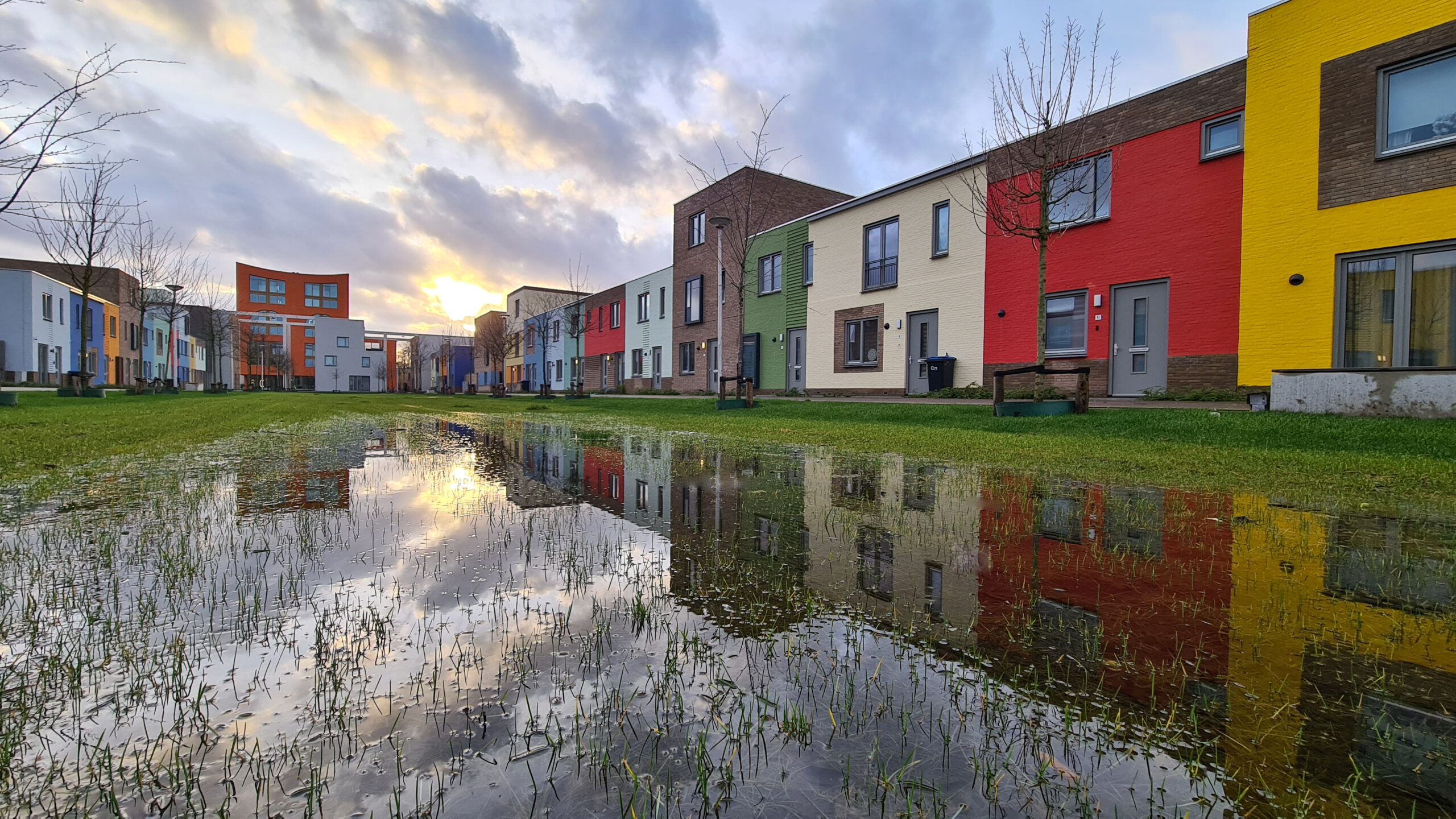 Rij kleurrijke huizen weerspiegeld in een grote plas, met bewolkte lucht en ondergaande zon.