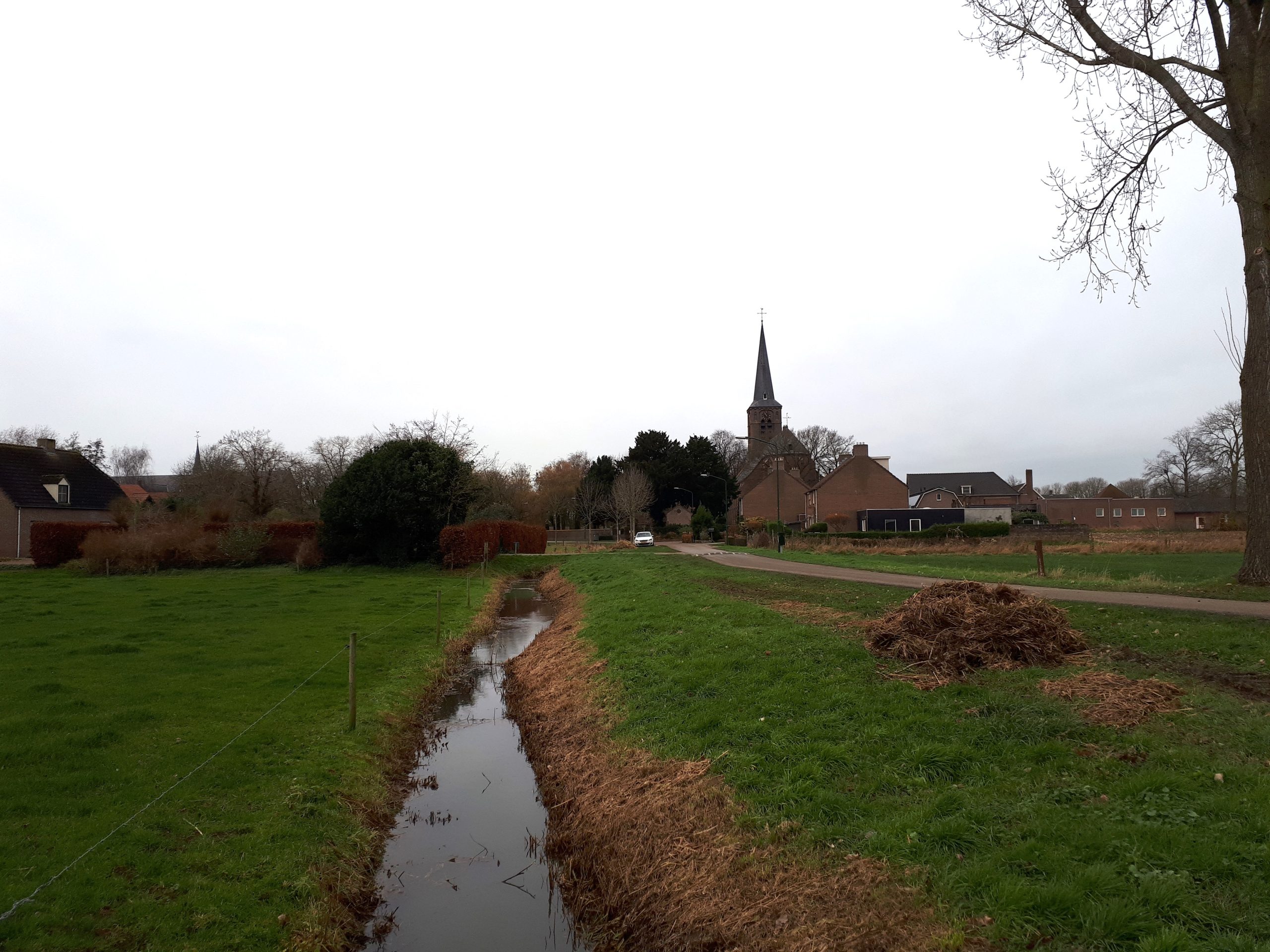 Platteland met grasveld, sloot, weg en kerk in de verte bij bewolkte hemel.